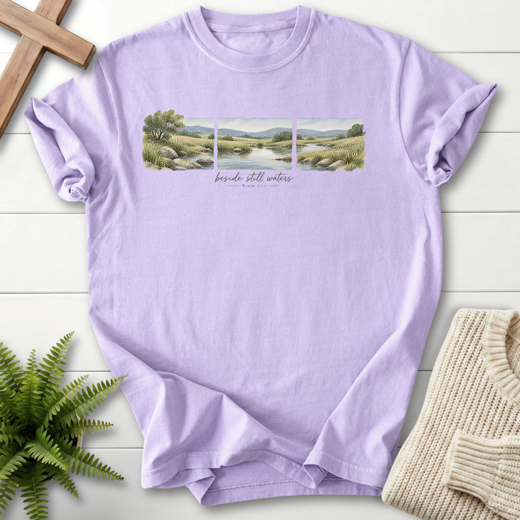 Flatlay of an Orchid Christian graphic tee with watercolor artwork with "Beside Still Waters T-Shirt" Psalm 23:2.