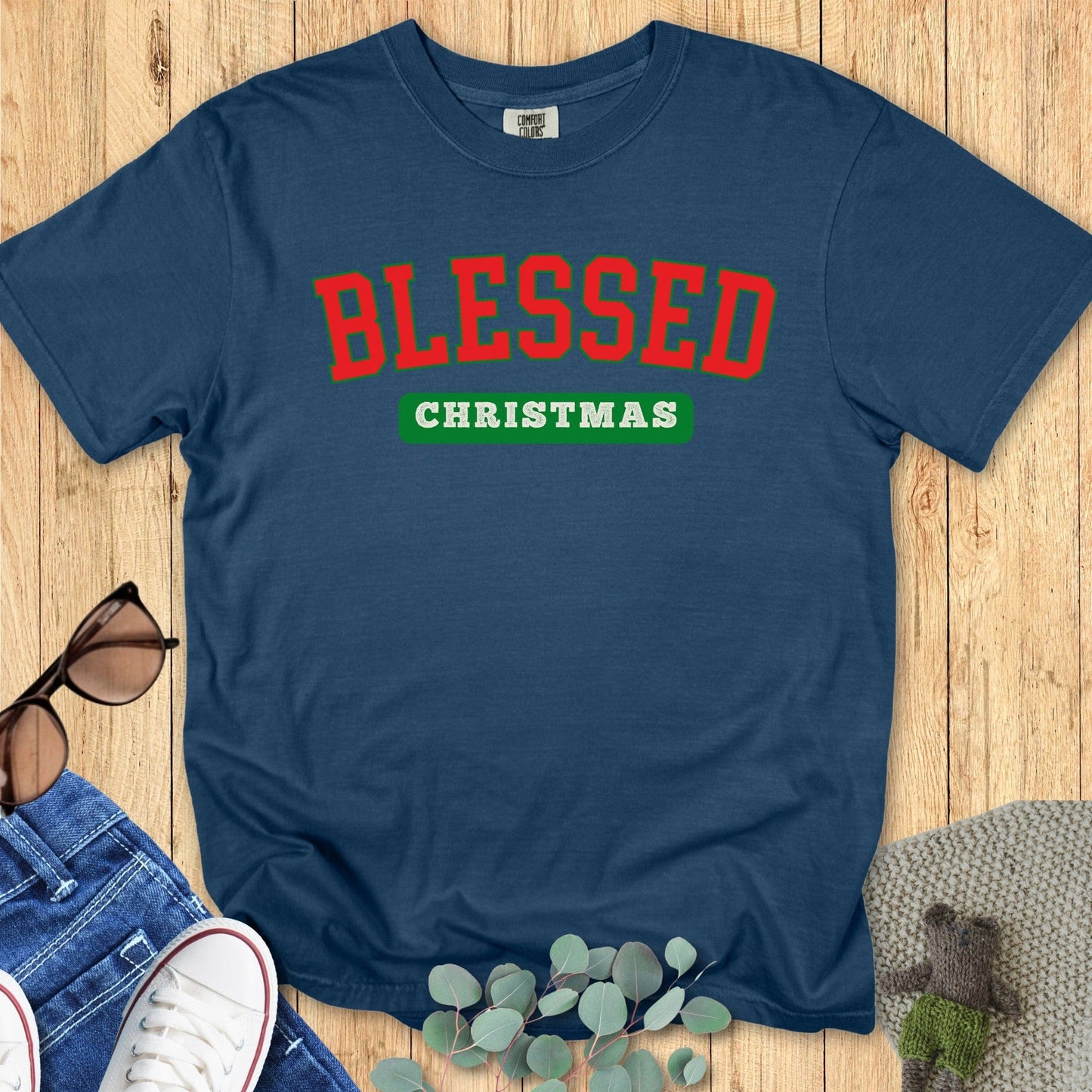Flatlay of a Comfort Colors 1717 midnight blue “Blessed Christmas” t-shirt with bold red and green varsity lettering. A festive women’s scripture tee — faith-based apparel for women and modern Christian shirt, thoughtful Christian mom gift idea for the holiday season.