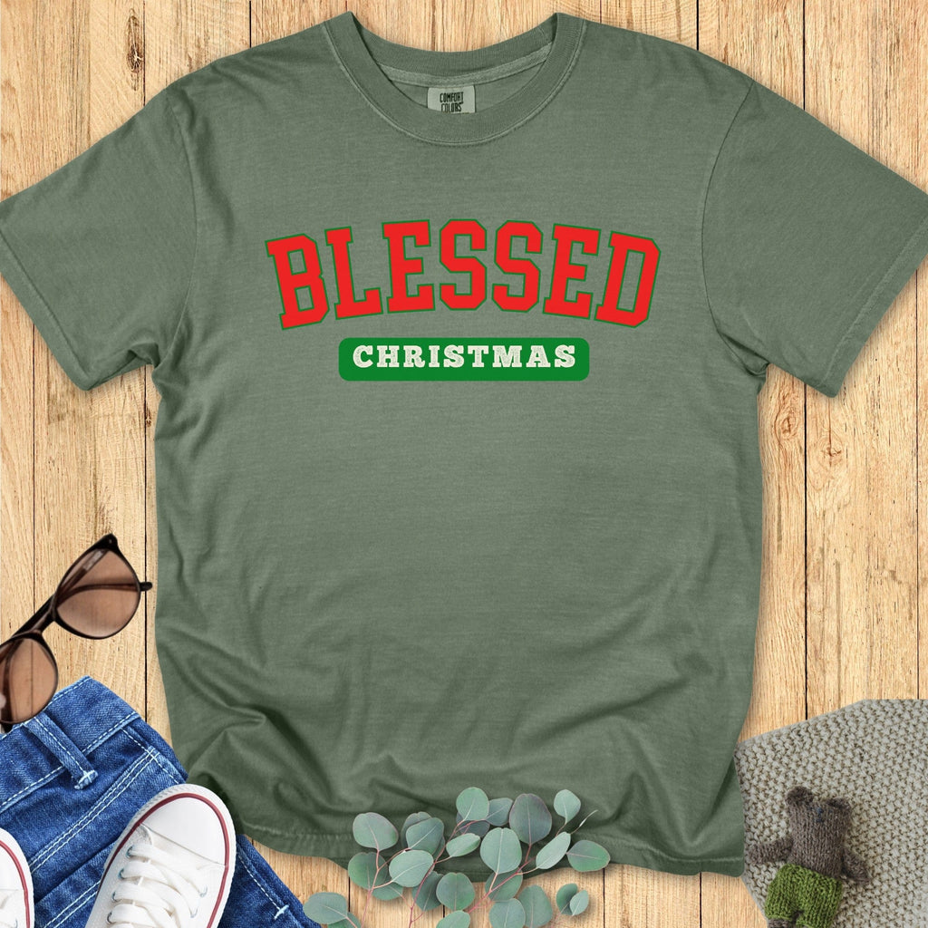 Flatlay of a Comfort Colors 1717 moss green “Blessed Christmas” t-shirt featuring classic red collegiate text and green accent. A cozy women’s scripture tee — faith-based apparel for women and modern Christian shirt, perfect Christian mom gift idea for Christmas gatherings.