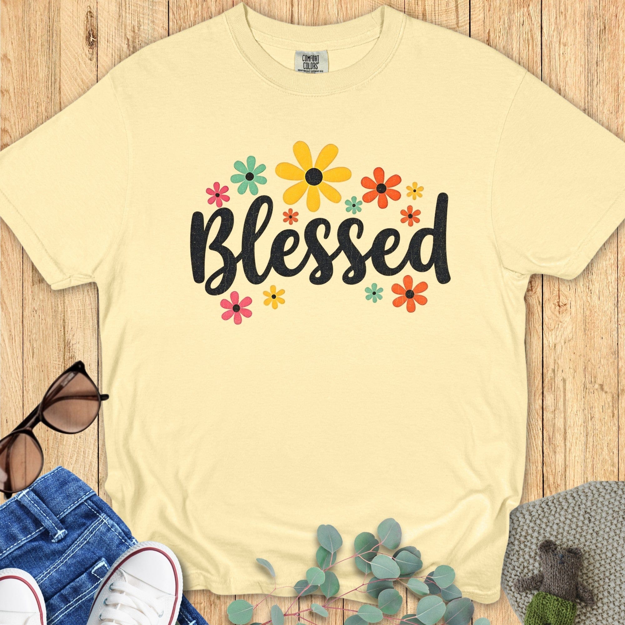 Flatlay of a Comfort Colors 1717 butter “Blessed” t-shirt with colorful floral graphics surrounding the word “Blessed.” A happy women’s scripture tee — faith-based apparel for women and modern Christian shirt, ideal Christian mom gift idea.