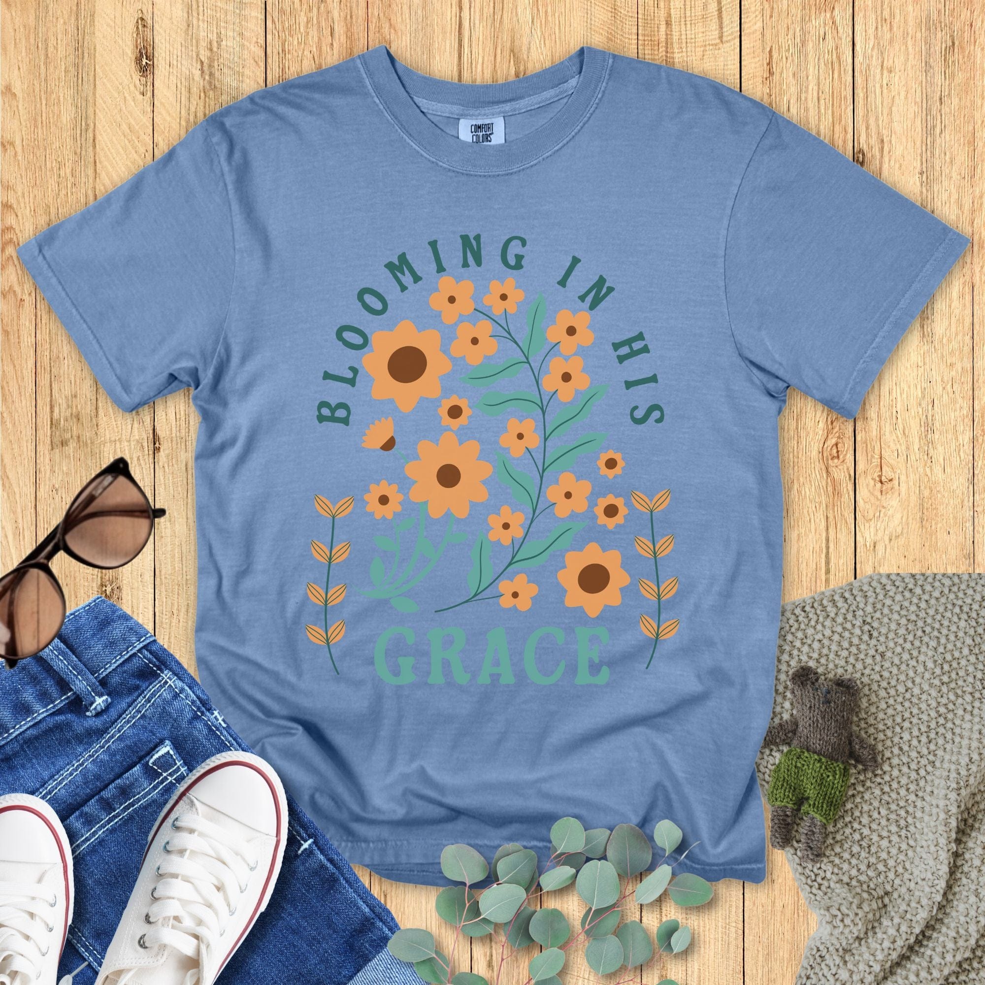Comfort Colors 1717 Christian women t-shirt in blue, printed with “Blooming in His Grace” in teal text surrounded by orange floral artwork. Styled flat with jeans, sneakers, sunglasses, eucalyptus sprigs, and a knitted bear on a wooden background.