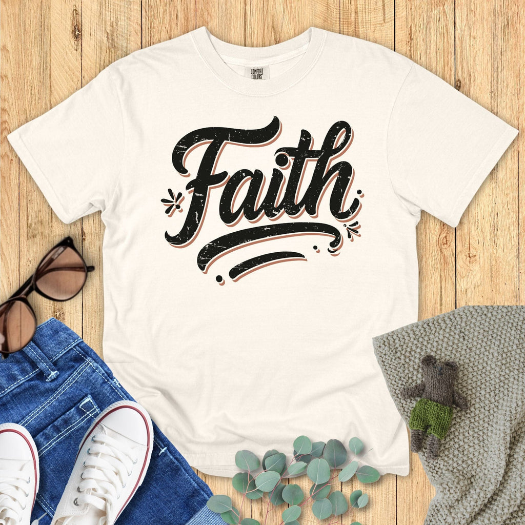 Flatlay of a Comfort Colors 1717 ivory “Faith” t-shirt featuring vintage black lettering, paired with denim and neutral accessories. A stylish piece from the modern Christian shirts collection, great for faith-based apparel for women.