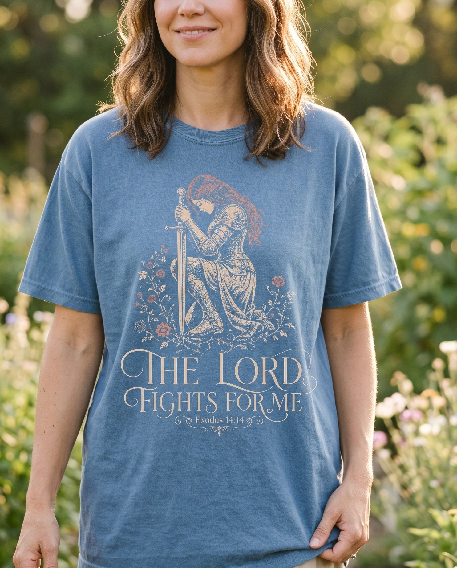 The LORD Fights For Me – Blue Jean women's Christian tee, lifestyle photo in outdoor garden – Female warrior in ornate armor kneeling in prayer with her sword plant – Exodus 14:14 – She Wears Faith