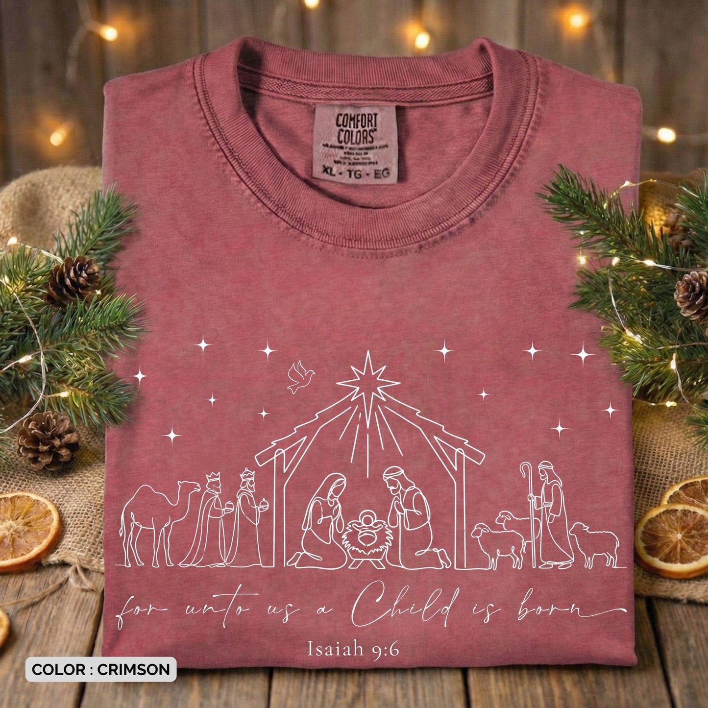 Folded Comfort Colors 1717 crimson Christian women’s T-shirt displaying a clean line-art nativity illustration and the scripture “For unto us a Child is born – Isaiah 9:6.” A festive holiday US faith wear tee set against rustic Christmas décor with pine branches and dried oranges.