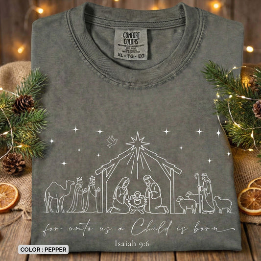 Folded Comfort Colors 1717 pepper Christian women tee featuring a minimalist nativity line-art design with Mary, Joseph, baby Jesus, the wise men, shepherds, and the Star of Bethlehem, with the verse “For unto us a Child is born – Isaiah 9:6.” A soft Christmas US faith wear T-shirt styled with holiday greenery and warm lights.