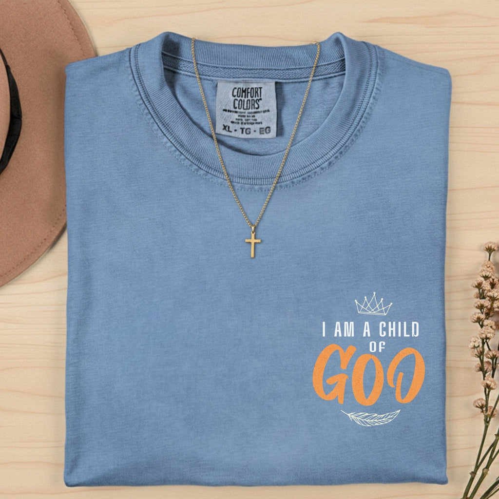 Comfort Colors 1717 blue jean “I Am a Child of God” t-shirt folded neatly with gold cross necklace and dried flowers. A classic women’s scripture tee — encouraging faith-based apparel for women and timeless modern Christian shirt, thoughtful Christian mom gift idea.