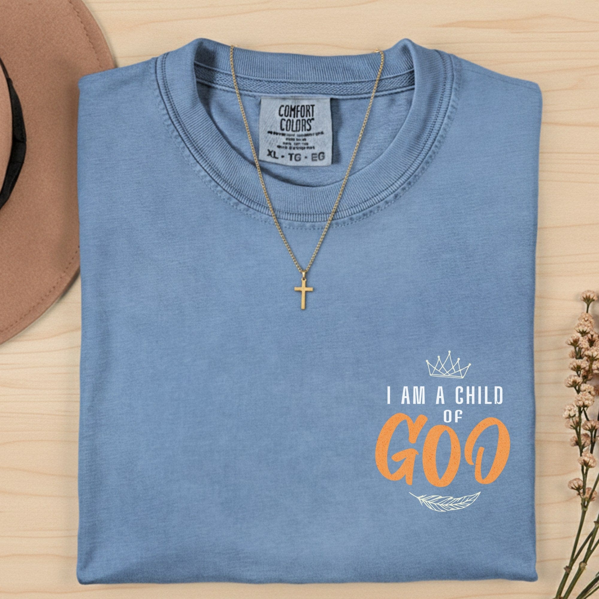 Comfort Colors 1717 blue jean “I Am a Child of God” t-shirt folded neatly with gold cross necklace and dried flowers. A classic women’s scripture tee — encouraging faith-based apparel for women and timeless modern Christian shirt, thoughtful Christian mom gift idea.
