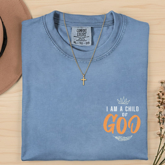 Comfort Colors 1717 blue jean “I Am a Child of God” t-shirt folded neatly with gold cross necklace and dried flowers. A classic women’s scripture tee — encouraging faith-based apparel for women and timeless modern Christian shirt, thoughtful Christian mom gift idea.