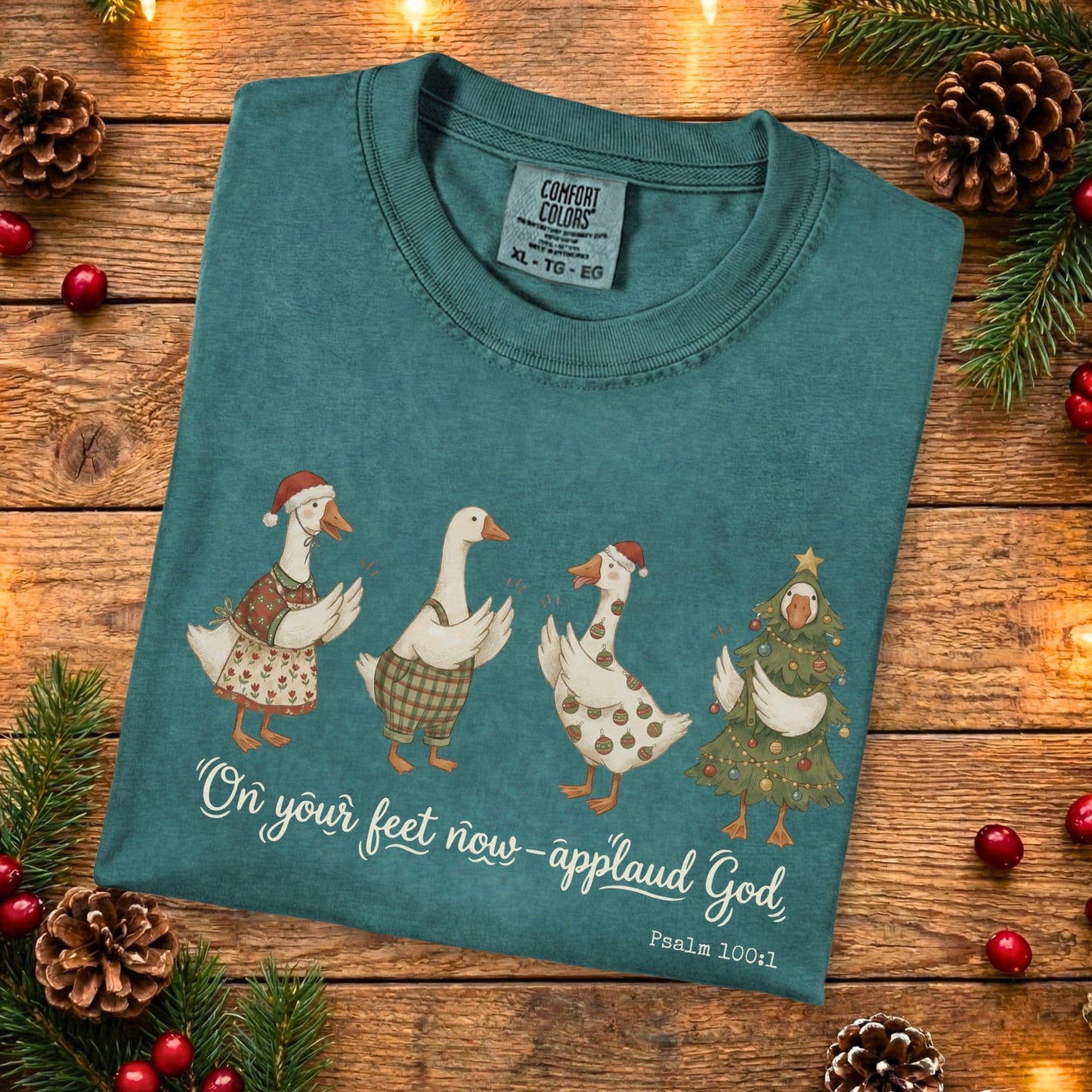 Folded Comfort Colors 1717 blue spruce Christian women tee featuring four festive geese dressed in Christmas outfits with the verse “On your feet now -applaud God, Psalm 100:1.” Cozy holiday US faith wear displayed on a rustic wooden background with pinecones and greenery.