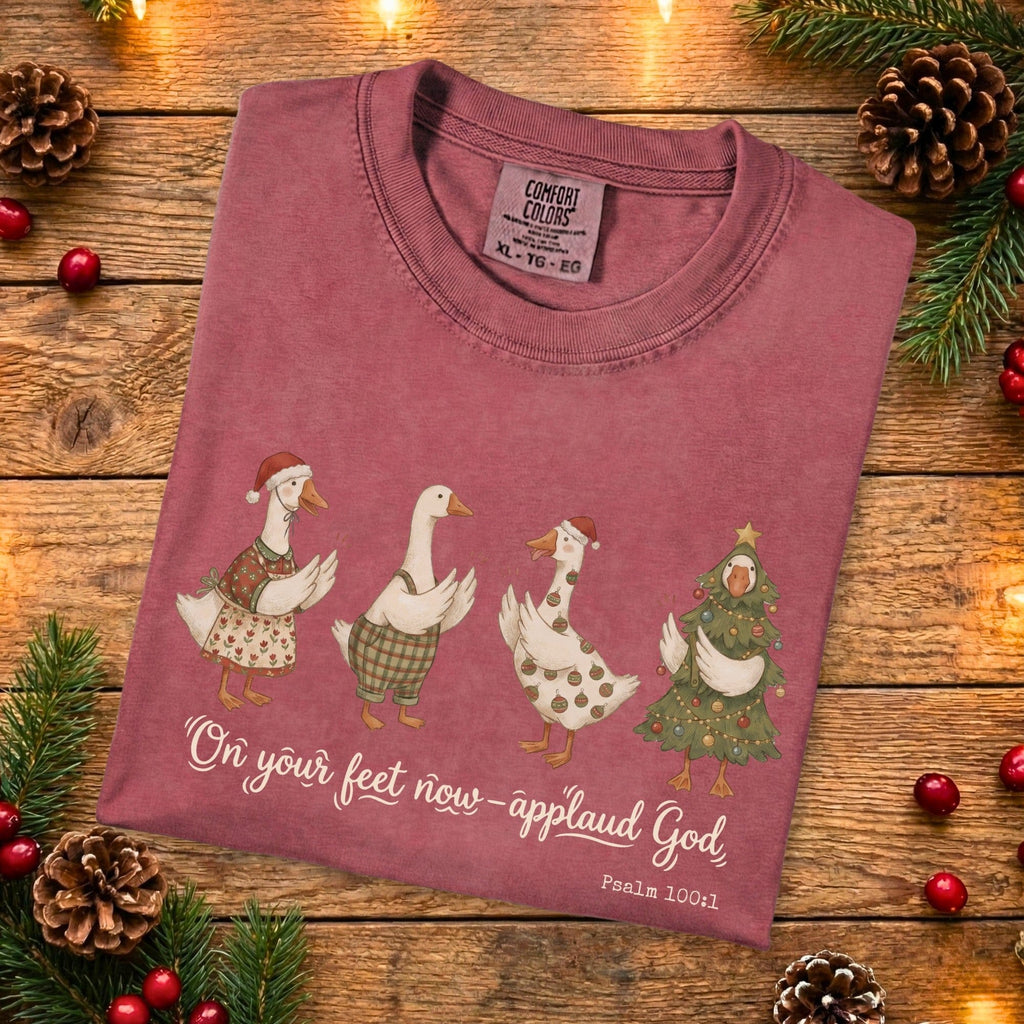 Folded Comfort Colors 1717 crimson Christian women’s T-shirt showing four whimsical Christmas geese and the scripture “On your feet now—applaud God, Psalm 100:1.” A soft, garment-dyed US faith wear holiday design styled with pinecones and Christmas decor.