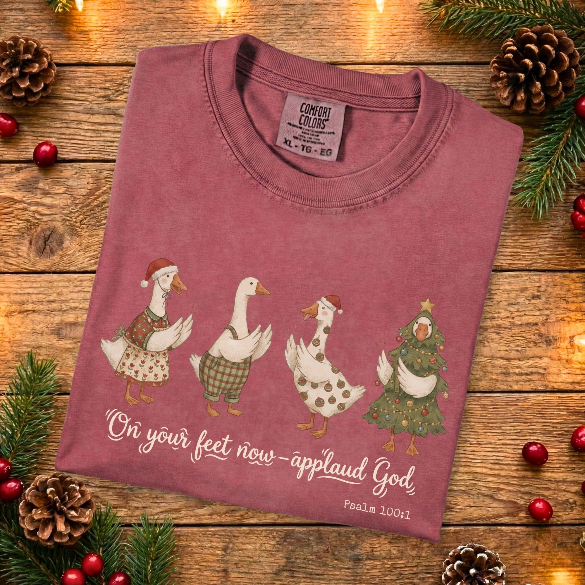 Folded Comfort Colors 1717 crimson Christian women’s T-shirt showing four whimsical Christmas geese and the scripture “On your feet now—applaud God, Psalm 100:1.” A soft, garment-dyed US faith wear holiday design styled with pinecones and Christmas decor.