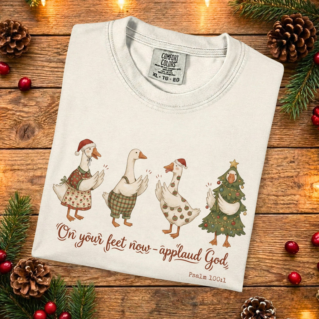 Folded Comfort Colors 1717 ivory Christian women tee with cute Christmas geese and the verse “On your feet now - applaud God, Psalm 100:1.” A festive US faith wear shirt photographed on a rustic wooden backdrop with pine branches and holiday accents.