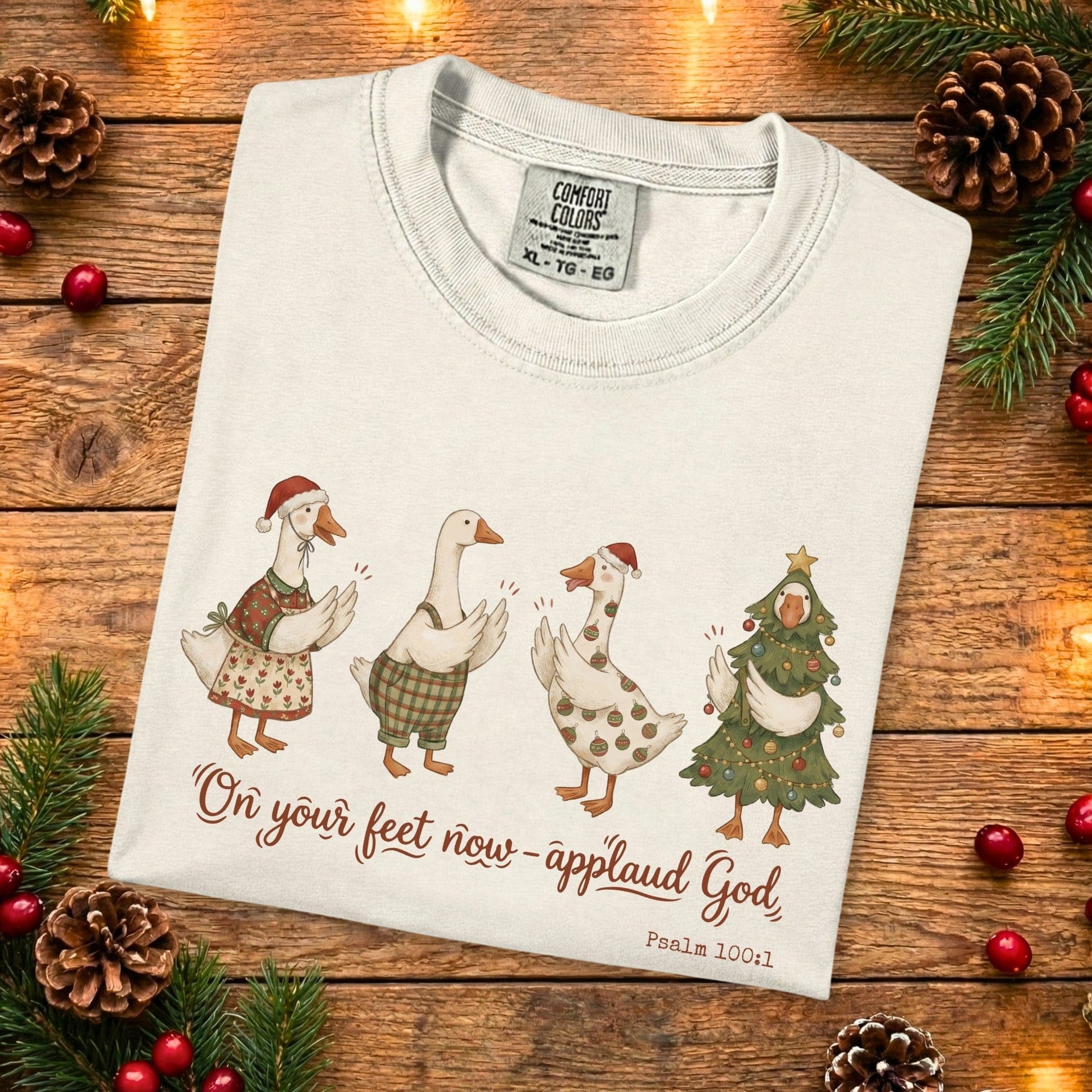 Folded Comfort Colors 1717 ivory Christian women tee with cute Christmas geese and the verse “On your feet now - applaud God, Psalm 100:1.” A festive US faith wear shirt photographed on a rustic wooden backdrop with pine branches and holiday accents.