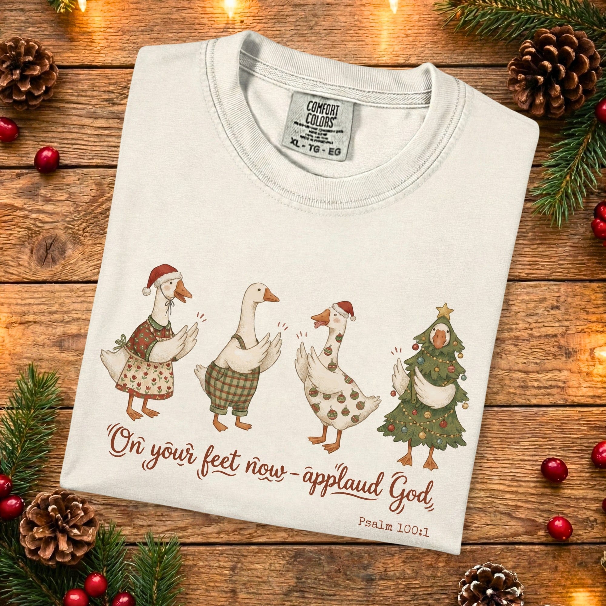 Folded Comfort Colors 1717 ivory Christian women tee with cute Christmas geese and the verse “On your feet now - applaud God, Psalm 100:1.” A festive US faith wear shirt photographed on a rustic wooden backdrop with pine branches and holiday accents.