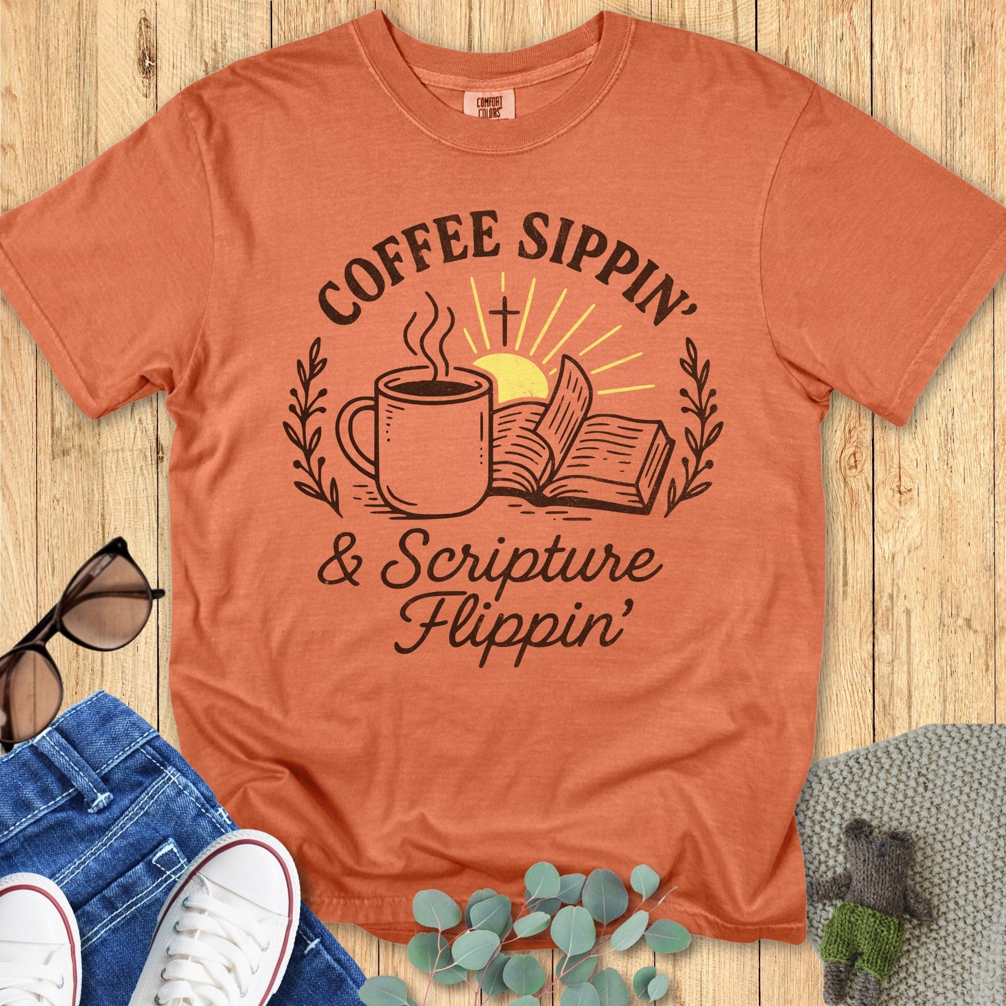 Flatlay of a Comfort Colors 1717 yam “Coffee Sippin’ & Scripture Flippin’” t-shirt with coffee cup and sunrise design. A warm-toned women’s scripture tee - faith-based apparel for women and modern Christian shirt, fun Christian mom gift idea for caffeine and scripture lovers.