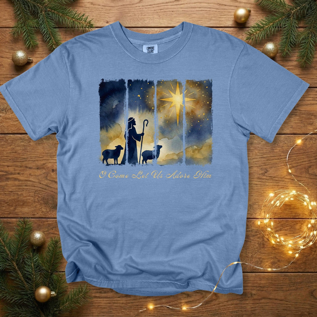 Comfort Colors 1717 blue jean Christian women tshirt showcasing a watercolor shepherd scene and bright Bethlehem star with “O Come Let Us Adore Him.” US faith wear tee styled on a rustic wood background with festive greenery and soft holiday lights.