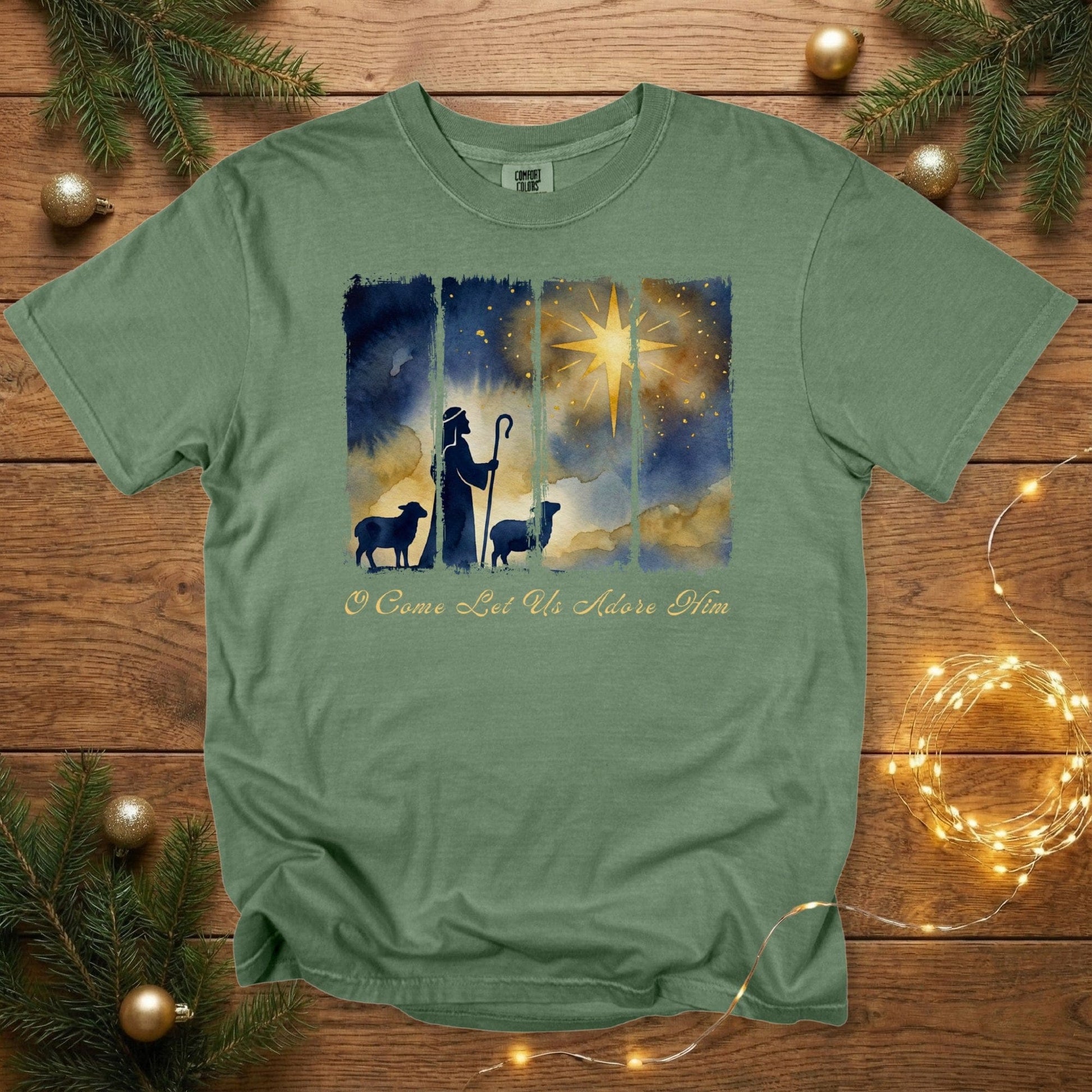 Comfort Colors 1717 hemp green Christian women tee featuring a watercolor shepherd silhouette, sheep, and radiant star above the words “O Come Let Us Adore Him.” Inspirational US faith wear tshirt placed on a wooden backdrop with Christmas greenery and twinkle lights.