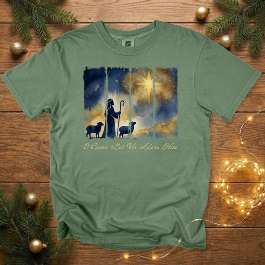 Comfort Colors 1717 hemp green Christian women tee featuring a watercolor shepherd silhouette, sheep, and radiant star above the words “O Come Let Us Adore Him.” Inspirational US faith wear tshirt placed on a wooden backdrop with Christmas greenery and twinkle lights.