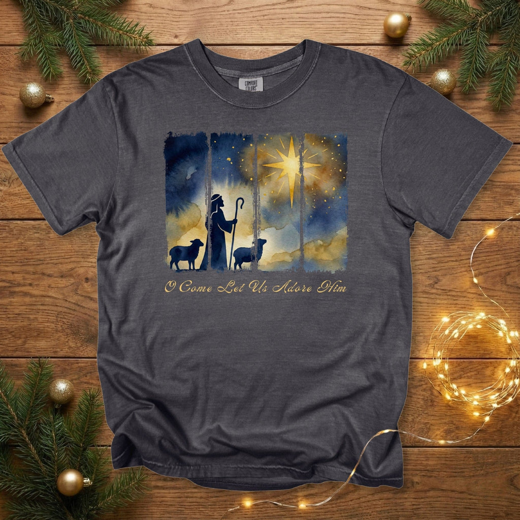 Comfort Colors 1717 pepper Christian women tee featuring a watercolor shepherd and sheep under the Christmas star with the phrase “O Come Let Us Adore Him.” Faith-based US Christian tshirt displayed on a wooden backdrop with Christmas greenery and warm lights.