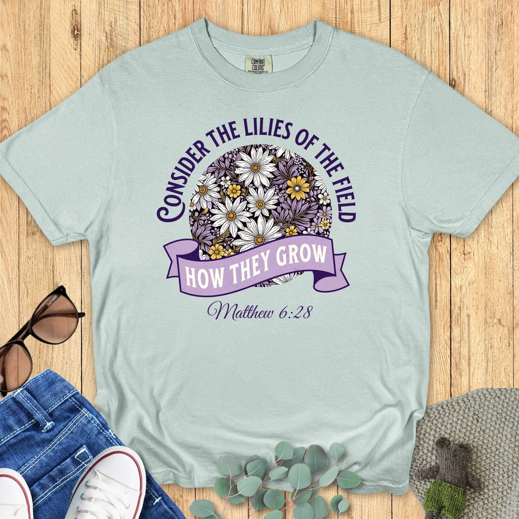 Comfort Colors 1717 Bay blue Christian T-shirt with scripture Matthew 6:28 and daisy illustration in purple, white, and yellow. Styled flat lay on wooden background with jeans and sneakers