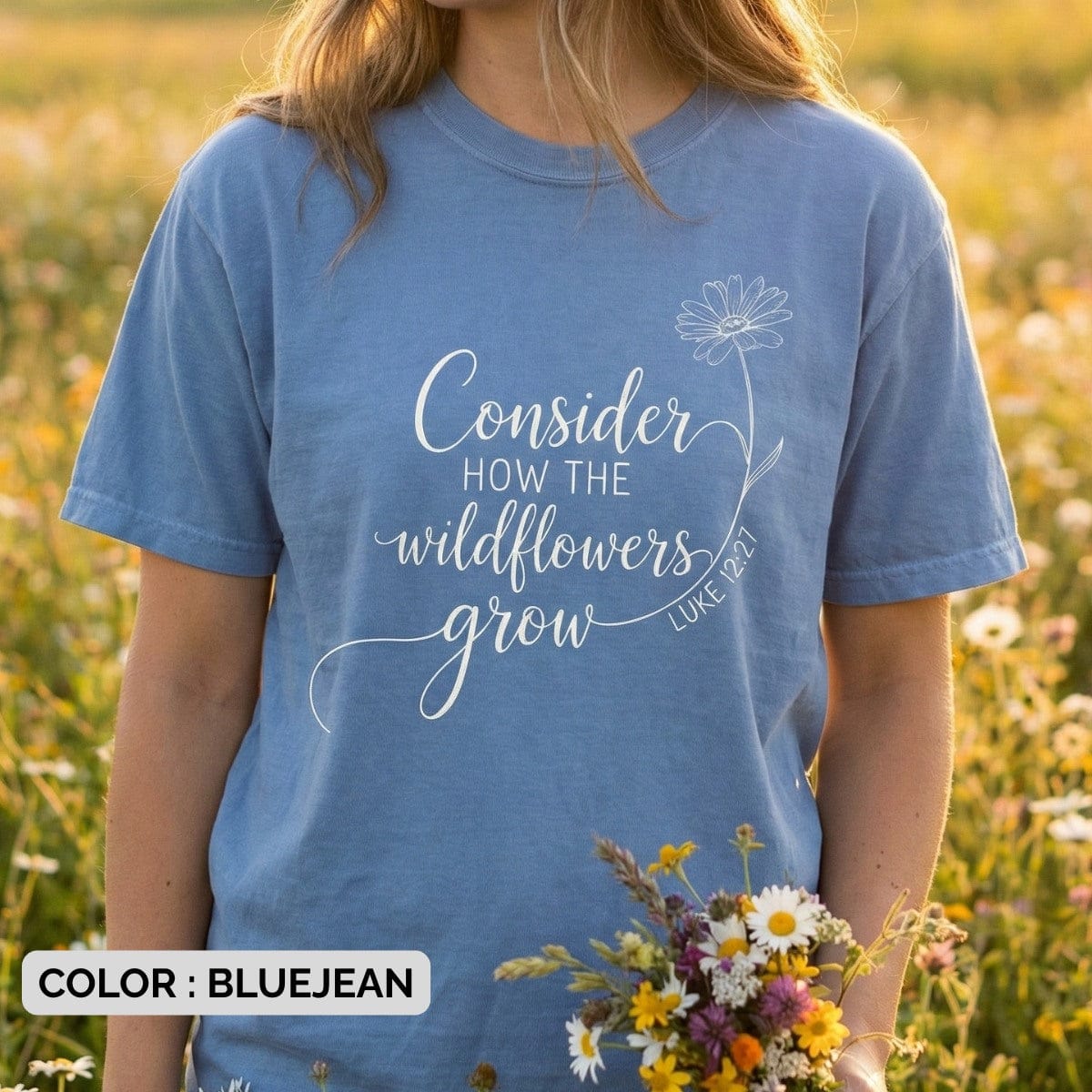 Woman standing in a flower field wearing a Blue Jean Comfort Colors 1717 tshirt with a white floral design. Lifestyle photography of a Christian women tee featuring "Consider how the wildflowers grow," showcasing natural US faith wear.