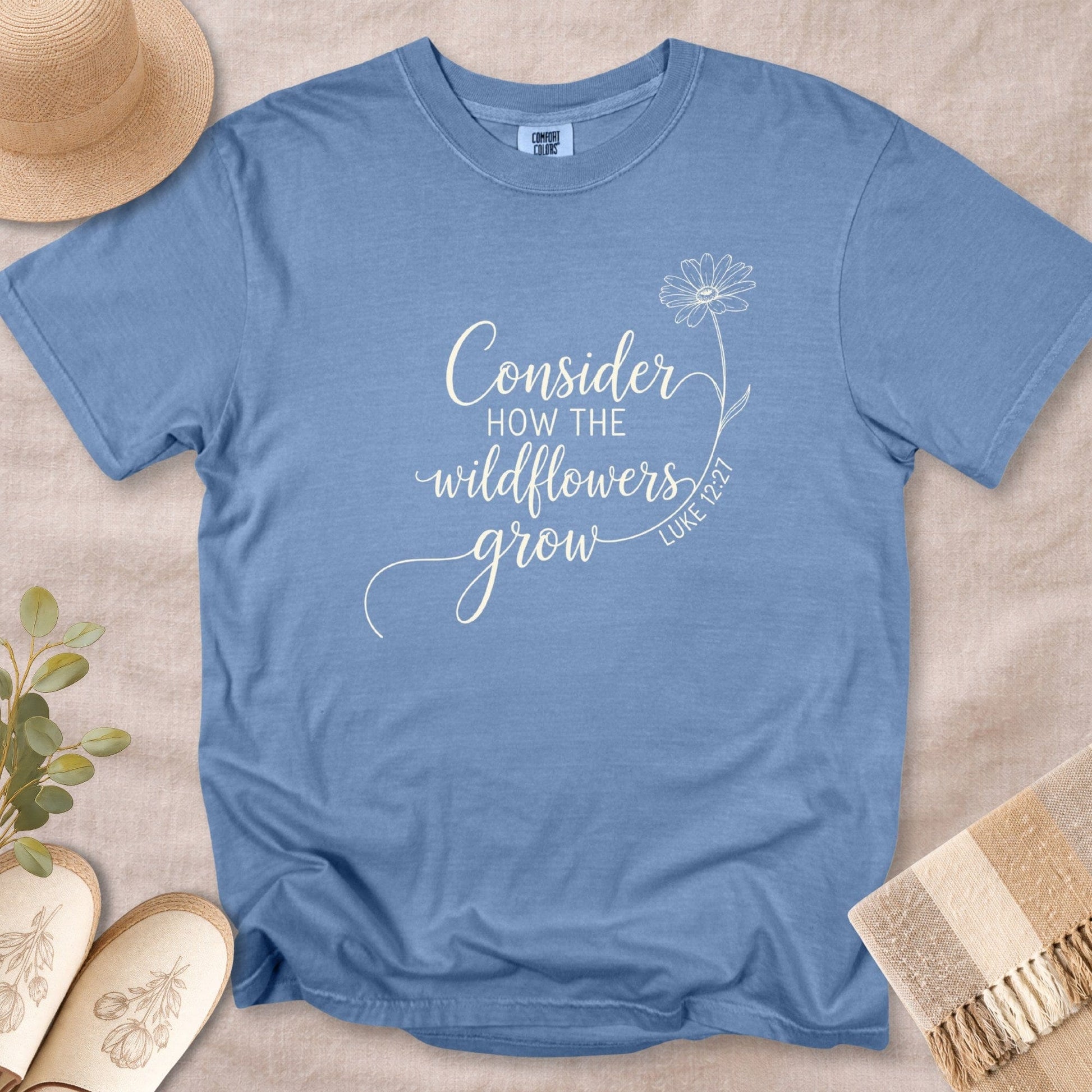 Blue Jean Comfort Colors 1717 tshirt with white calligraphy and a flower drawing. A soft, garment-dyed Christian women tee featuring Luke 12:27, a popular choice for US faith wear.