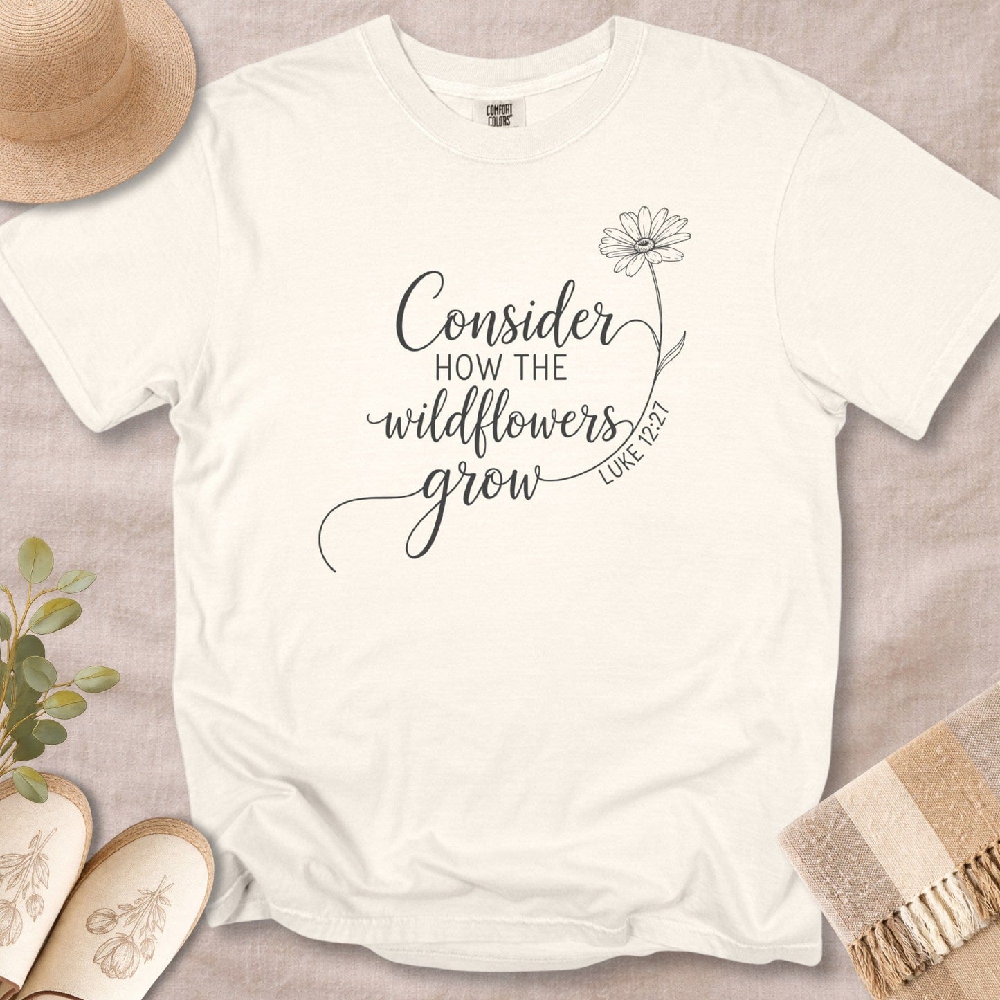 Flat lay of an Ivory Comfort Colors 1717 tshirt featuring dark grey text and a wildflower sketch. A vintage-inspired Christian women tee with the Luke 12:27 Bible verse, styled for US faith wear shops.