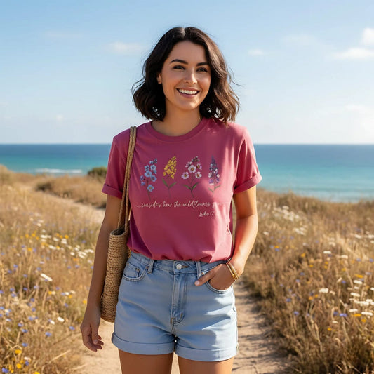 A smiling woman by the seaside wearing a Comfort Colors 1717 Christian women t-shirt in crimson. The shirt features colorful wildflower illustrations with the Bible verse “Consider how the wildflowers grow – Luke 12:27” in script font. The relaxed fit and soft vintage style highlight the faith-based design