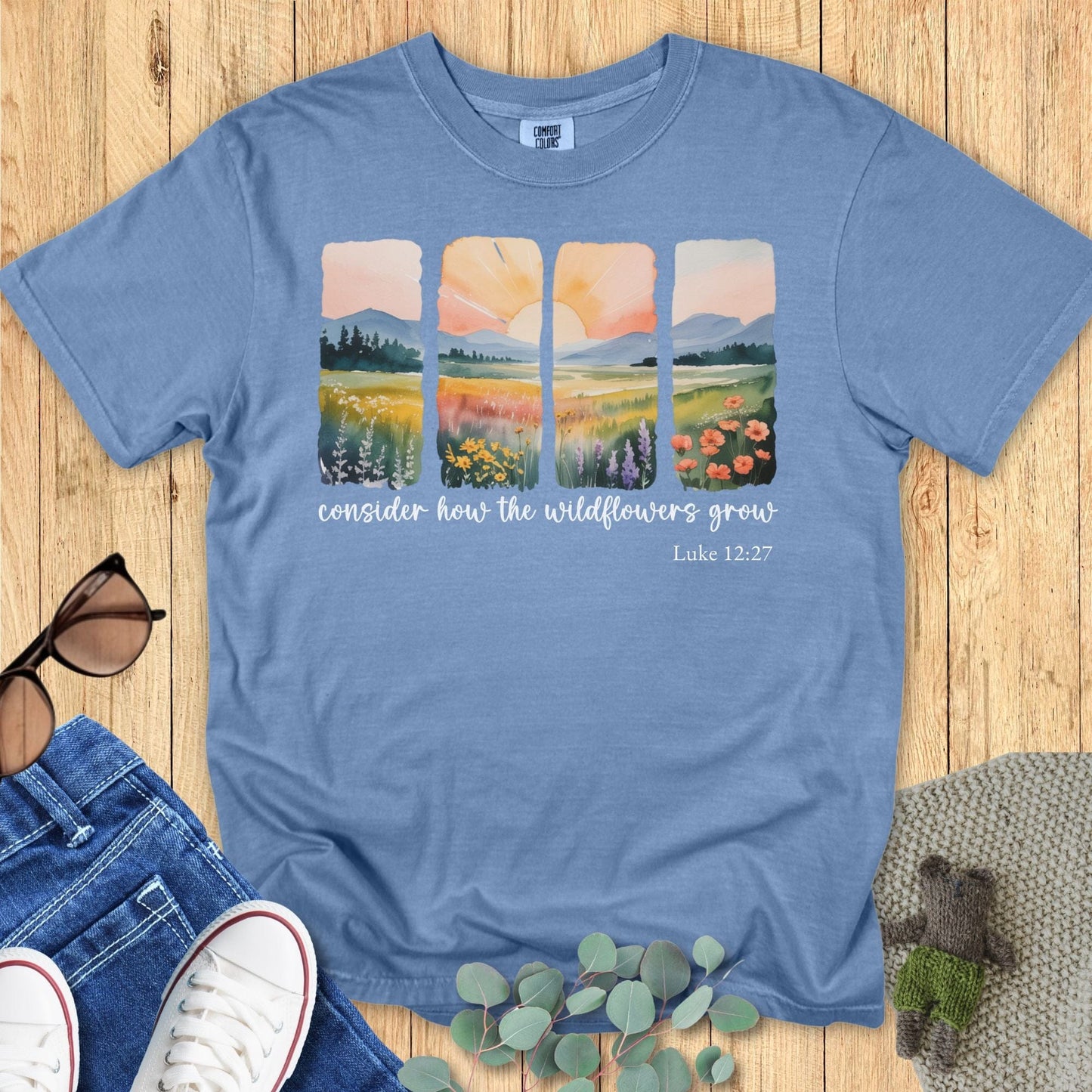 Blue Jean Comfort Colors T-shirt featuring watercolor-style four-panel wildflower landscape and sunrise design, scripture “Consider how the wildflowers grow – Luke 12:27.” Flat lay with jeans, sneakers, sunglasses, and eucalyptus on wooden background