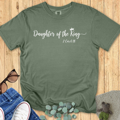 Moss green Comfort Colors T-shirt with the phrase “Daughter of the King” in elegant white script with a small crown graphic, featuring 2 Corinthians 6:18 beneath. Flat lay style with jeans, white sneakers, sunglasses, and eucalyptus leaves on a wooden background