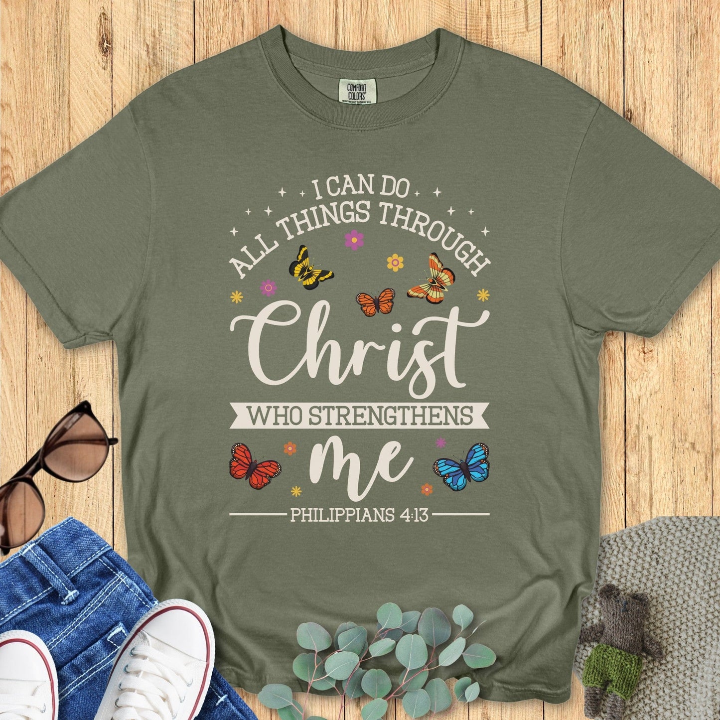 Flatlay of a Comfort Colors 1717 sage “I Can Do All Things Through Christ Who Strengthens Me – Philippians 4:13” t-shirt featuring colorful butterflies and floral accents. A joyful women’s scripture tee and uplifting faith-based apparel for women.