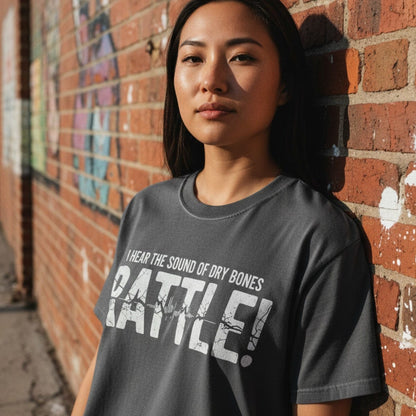 Woman standing by a brick wall wearing a Comfort Colors 1717 pepper “I Hear the Sound of Dry Bones RATTLE!” t-shirt. A bold women’s scripture tee — powerful faith-based apparel for women and modern Christian shirt, unique Christian mom gift idea inspired by Ezekiel 37.
