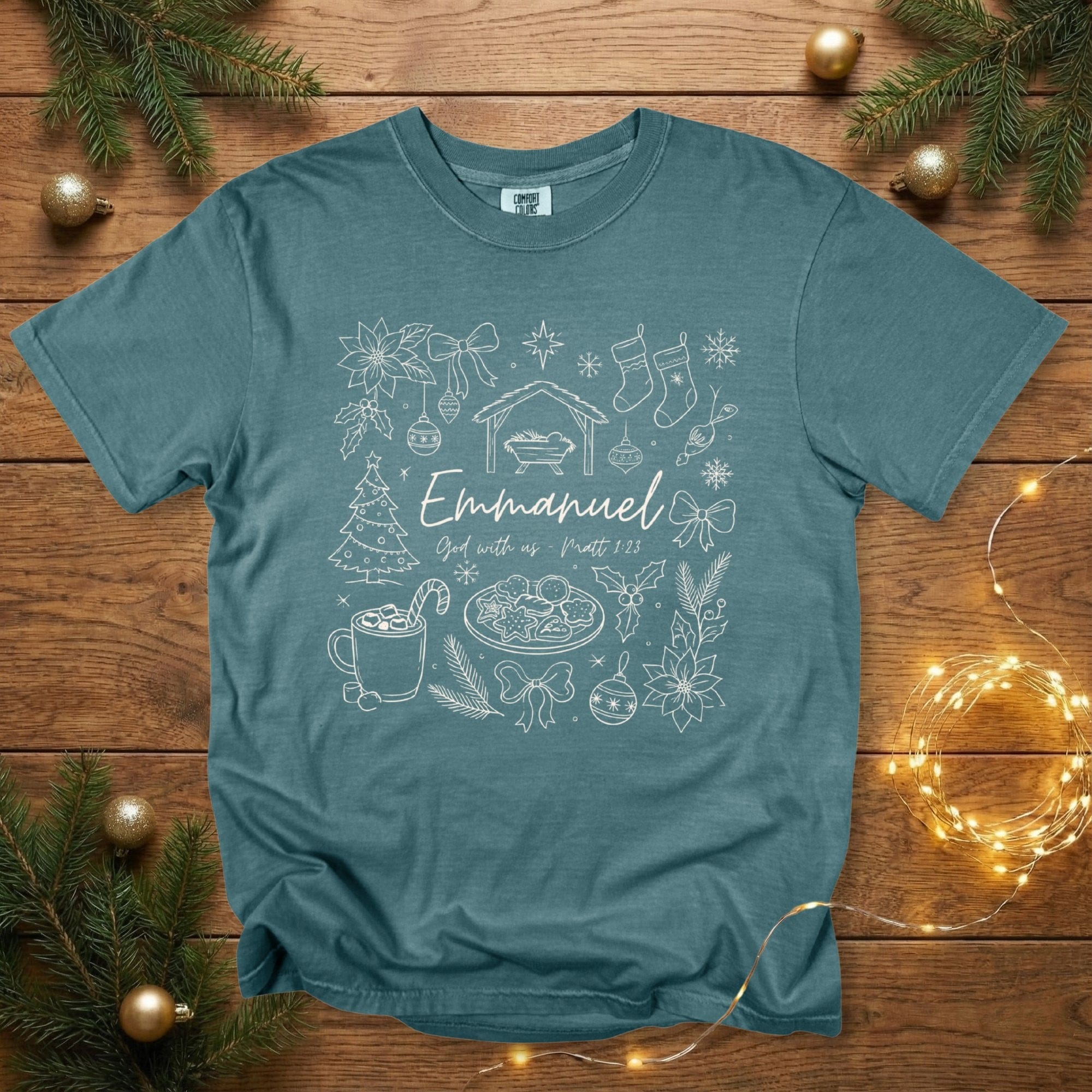 Comfort Colors 1717 blue spruce Christian women tee showcasing a whimsical Christmas line-art design featuring the nativity scene, festive decorations, cookies, bows, and the words “Emmanuel – God with us, Matt 1:23.” A cozy, scripture-rooted US faith wear shirt for Christmas.