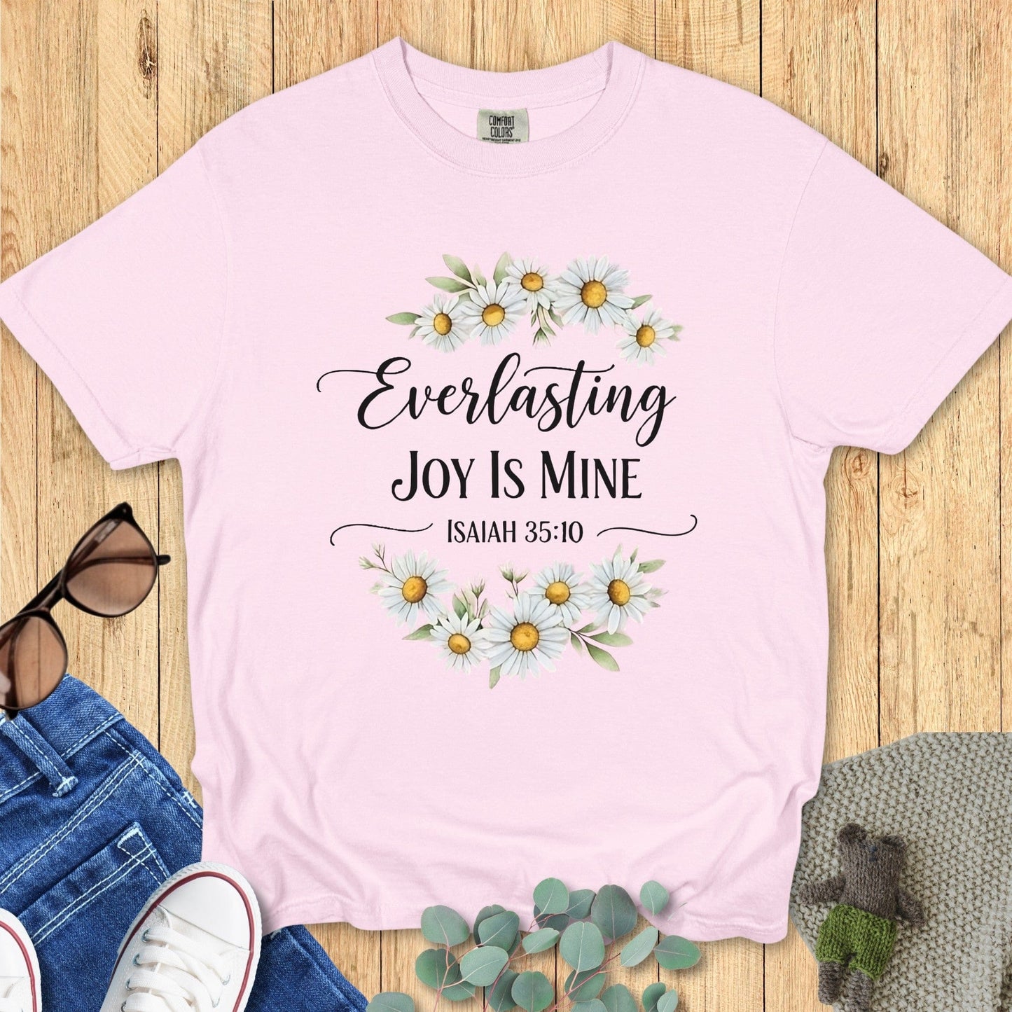 Flatlay of a Comfort Colors 1717 blossom pink “Everlasting Joy Is Mine – Isaiah 35:10” t-shirt with soft floral accents. A gentle and feminine addition to modern Christian shirts collection.