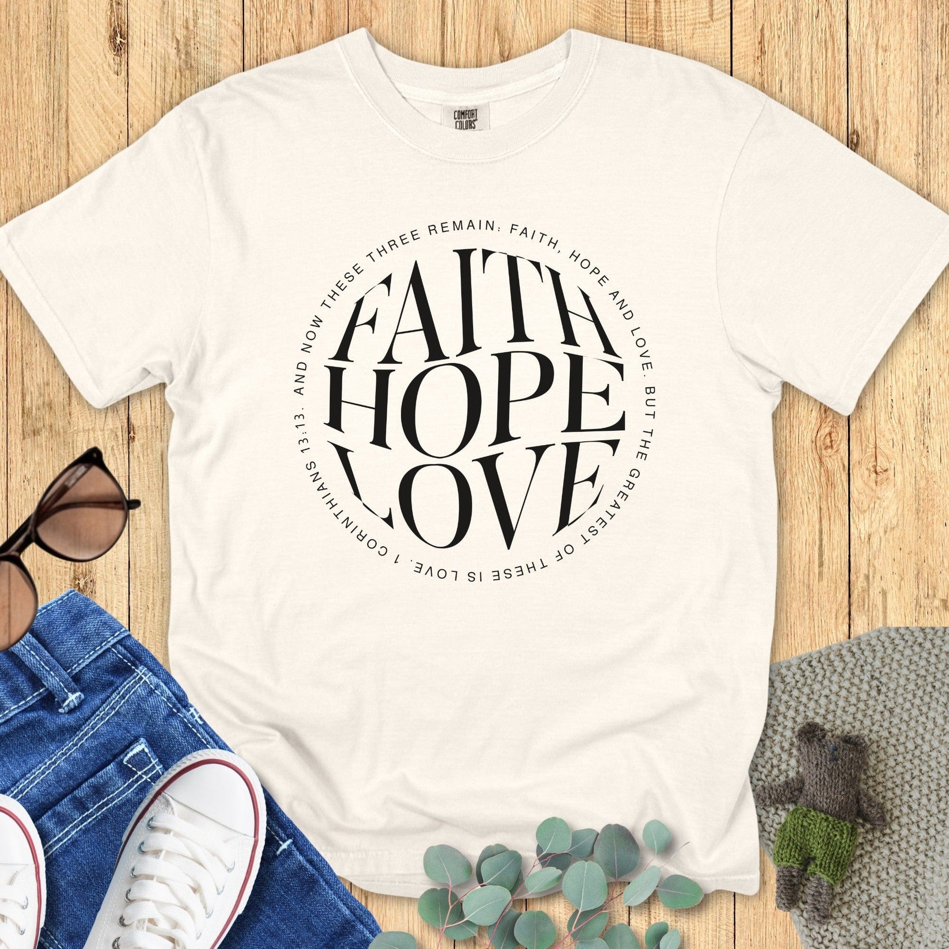 latlay of a Comfort Colors 1717 ivory “Faith Hope Love” t-shirt featuring 1 Corinthians 13:13 in elegant circular text. A timeless women’s scripture tee -  faith-based apparel for women and modern Christian shirt, heartfelt Christian mom gift idea reflecting God’s enduring love.