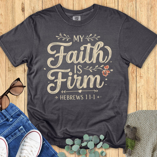 Comfort Colors 1717 pepper black Christian women’s t-shirt featuring the inspirational verse ‘My Faith Is Firm – Hebrews 11:1’ in vintage cream lettering with small floral details, displayed in lifestyle flat lay with casual accessories