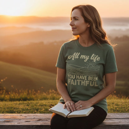 Woman sitting on a bench at sunset with an open Bible, wearing a moss green Comfort Colors 1717 Christian women t-shirt. The shirt design reads “All my life you have been faithful” with a sunrise line art graphic, symbolizing faith and God’s goodness