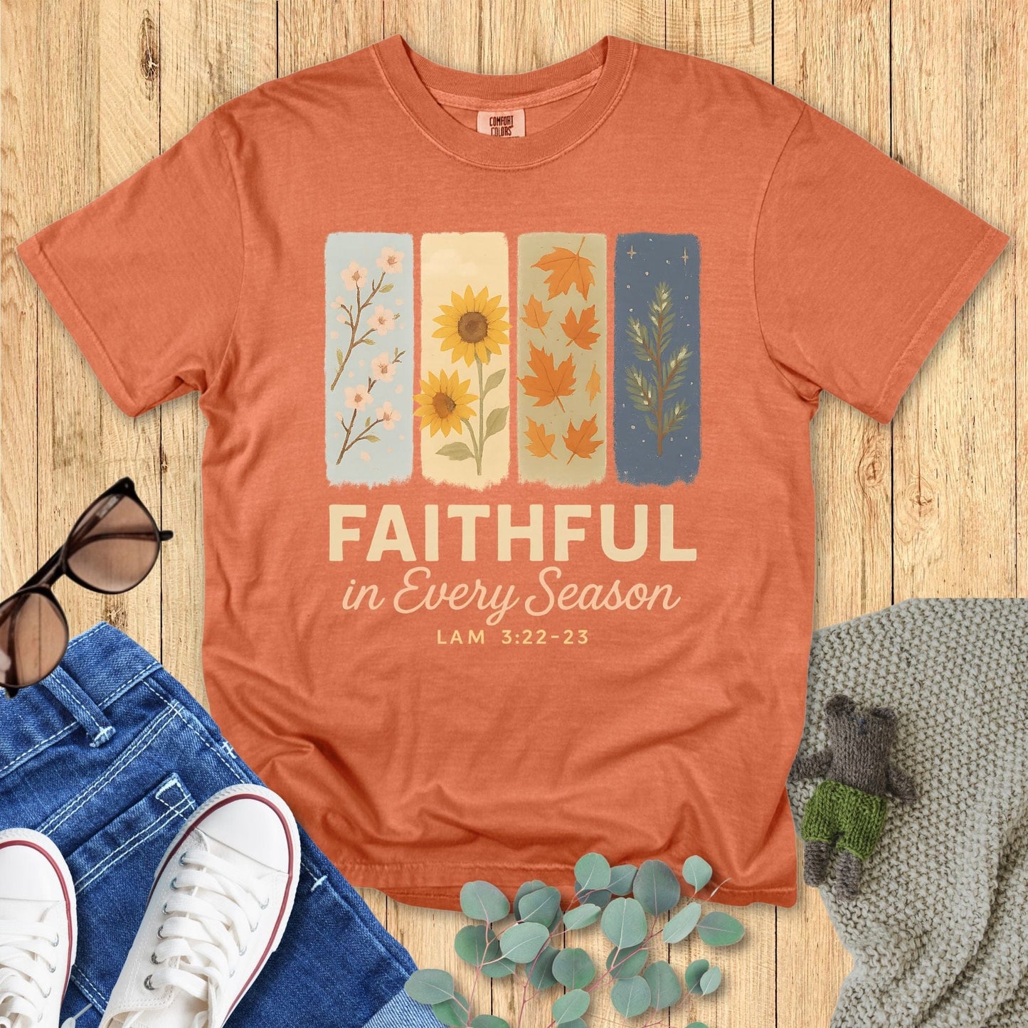 Flat lay of Comfort Colors 1717 Terracotta Christian women’s t-shirt with the text “Faithful in Every Season – Lam 3:22-23.” Design features four illustrated seasonal panels with pastel backgrounds. Styled with denim jeans, white sneakers, sunglasses, and eucalyptus leaves on a wooden backdro