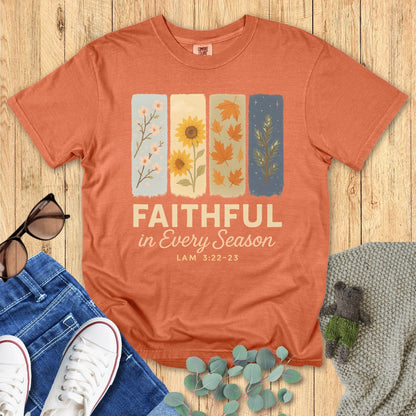 Flat lay of Comfort Colors 1717 Terracotta Christian women’s t-shirt with the text “Faithful in Every Season – Lam 3:22-23.” Design features four illustrated seasonal panels with pastel backgrounds. Styled with denim jeans, white sneakers, sunglasses, and eucalyptus leaves on a wooden backdro