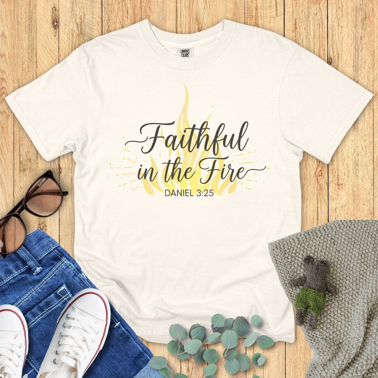 White t-shirt with 'Faithful in the Fire' text on a wooden surface with accessories.