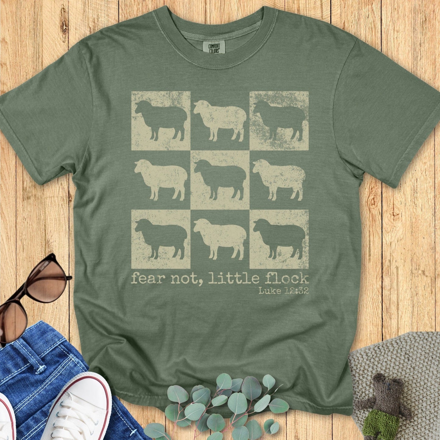 Comfort Colors 1717 Christian women t-shirt in moss green featuring a 3x3 sheep grid design with distressed texture and the Bible verse text "fear not, little flock – Luke 12:32," styled on a wooden background with casual accessories.