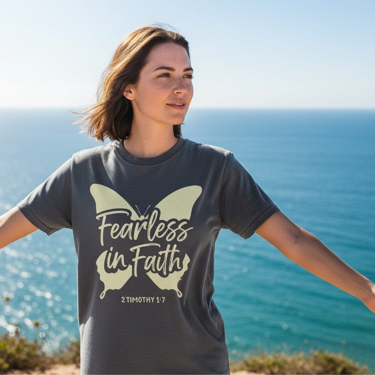 Woman standing by the ocean wearing a Comfort Colors 1717 pepper “Fearless in Faith – 2 Timothy 1:7” t-shirt with a butterfly design. A bold women’s scripture tee — empowering faith-based apparel for women and modern Christian shirt, meaningful Christian mom gift idea.