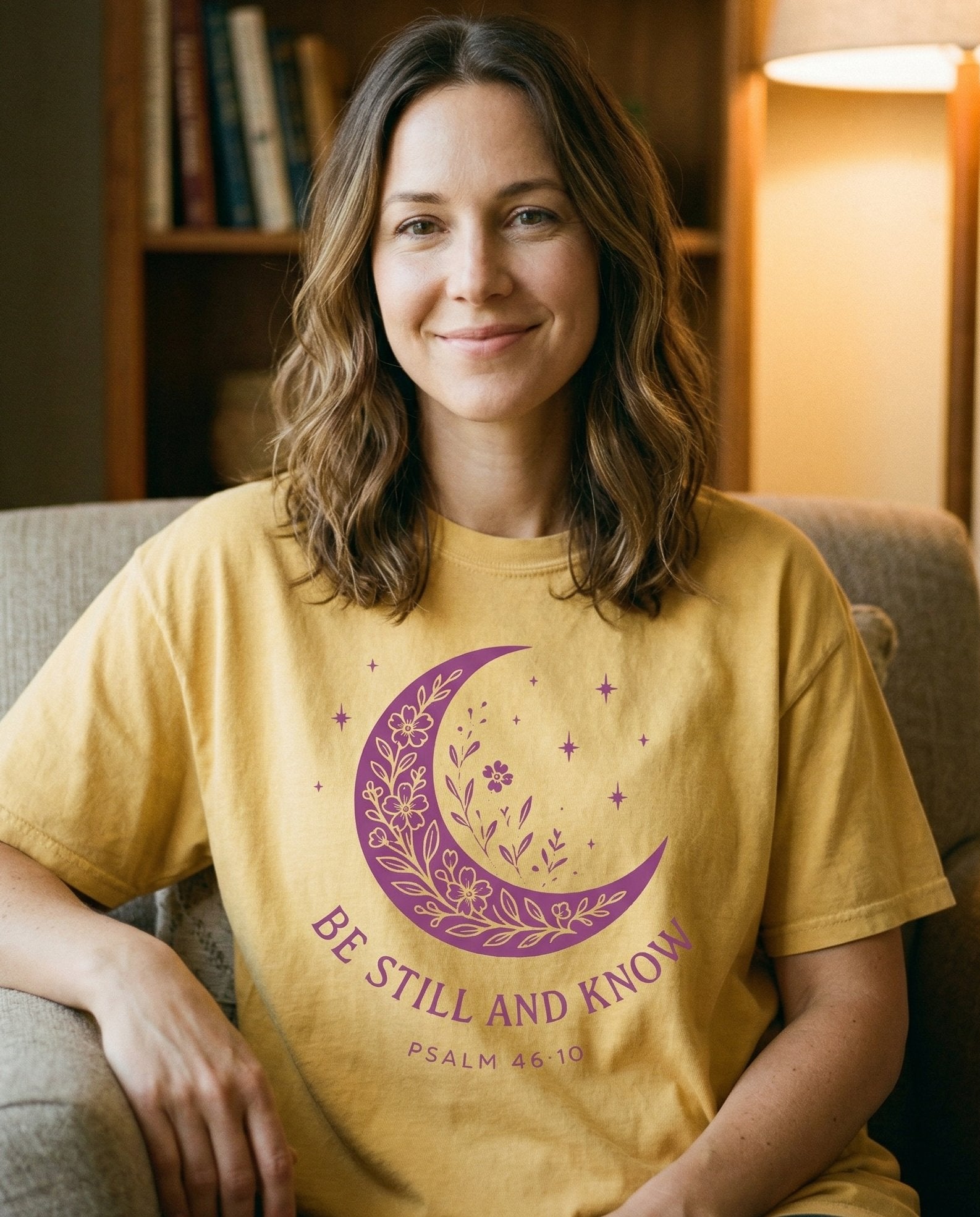 Be Still and Know Floral Moon – Butter women's Christian tee, lifestyle photo in cozy reading nook – Purple crescent moon filled with wildflowers and botanical motifs surr – Psalm 46:10 – She Wears Faith
