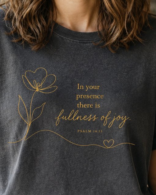 Fullness of Joy Dark - Pepper women's Christian tee, lifestyle photo in cozy café - Gold line-art flower on charcoal - Psalm 16:11 - She Wears Faith