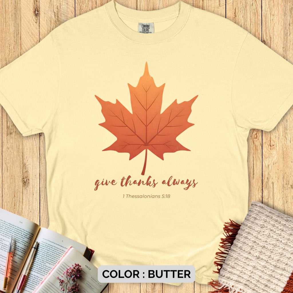 Faith-based Comfort Colors 1717 tee with maple leaf and “Give Thanks Always” Bible verse design, displayed flat on wood background with open Bible and cozy autumn props.
