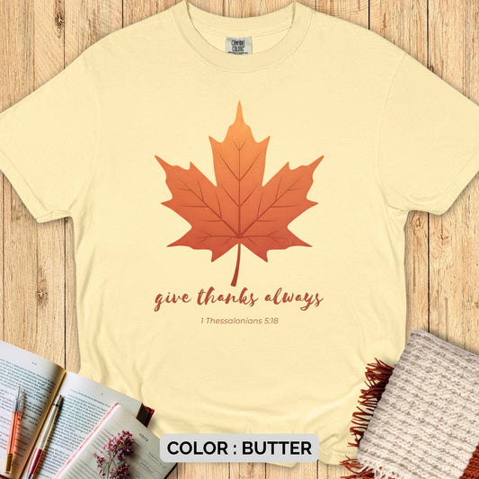 Faith-based Comfort Colors 1717 tee with maple leaf and “Give Thanks Always” Bible verse design, displayed flat on wood background with open Bible and cozy autumn props.
