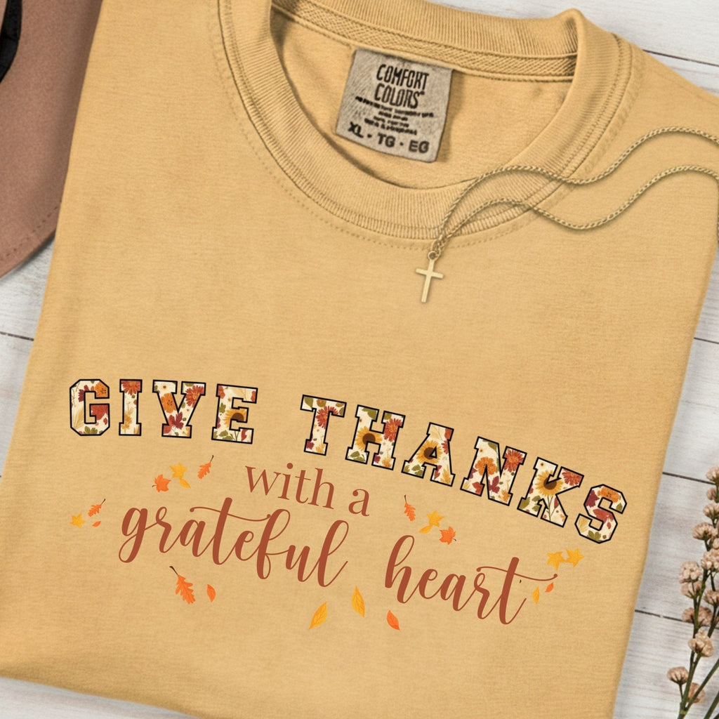 Folded Comfort Colors 1717 mustard “Give Thanks with a Grateful Heart” t-shirt featuring fall floral lettering and autumn leaves. A cozy women’s scripture tee - faith-based apparel for women and modern Christian shirt, perfect Christian mom gift idea for Thanksgiving or everyday gratitude.