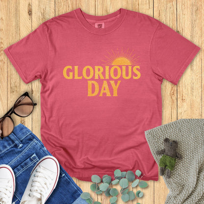 Flatlay of a Comfort Colors 1717 crimson “Glorious Day” t-shirt with cheerful golden typography and retro sunrise detail. A bright women’s scripture tee — encouraging faith-based apparel for women and modern Christian shirt, perfect Christian mom gift idea.