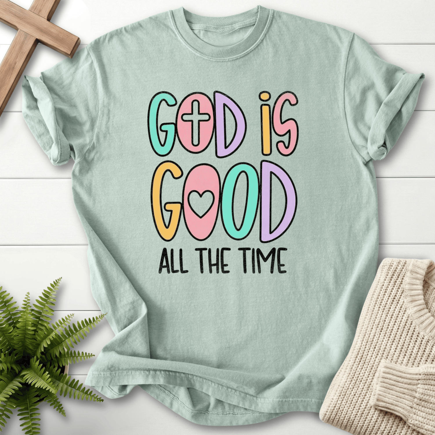 Bay blue women's faith t-shirt with bright multicolor 'God Is Good' lettering and a cross motif.