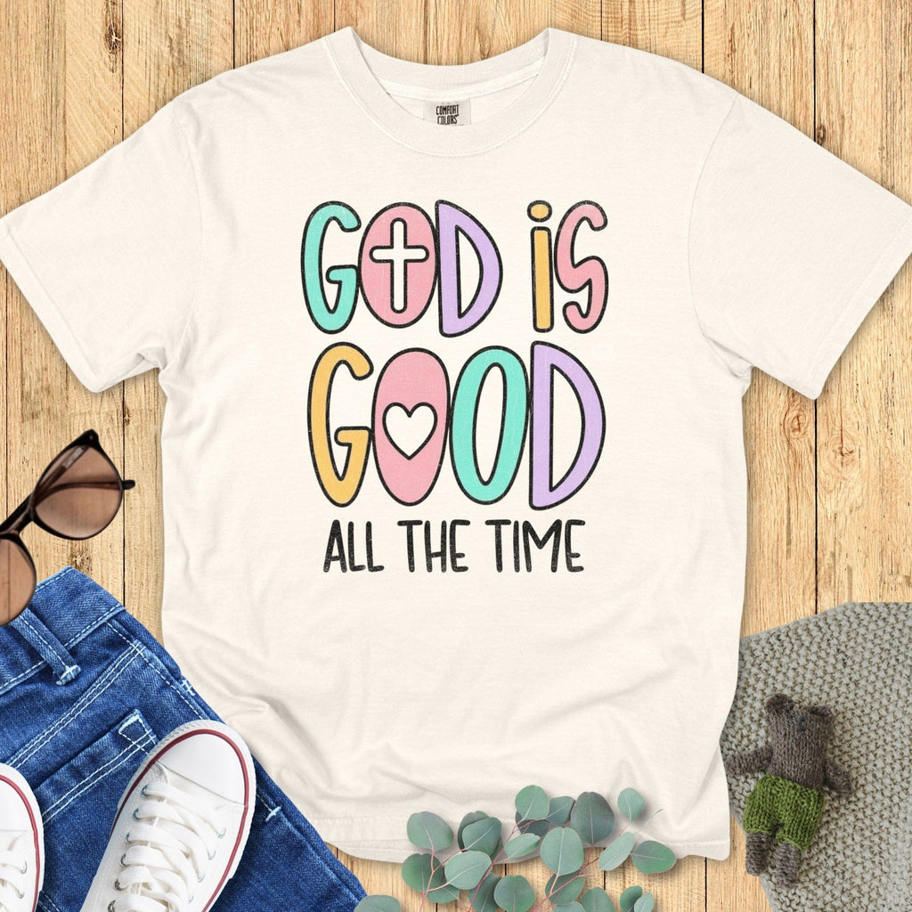 Flatlay of a Comfort Colors 1717 ivory “God Is Good All The Time” t-shirt with playful pastel typography and cross and heart icons. A cheerful women’s scripture tee - faith-based apparel for women and modern Christian shirt, uplifting Christian mom gift idea for everyday encouragement.